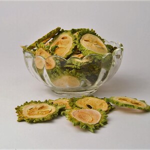 May include: A clear glass bowl filled with sliced, dried bitter melon. The slices are green and yellow, with a textured, ridged exterior. Some slices are scattered around the bowl on a white surface.