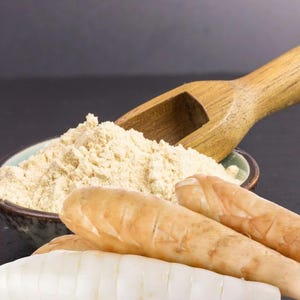 Puede incluir: Primer plano de un cuenco lleno de polvo marrón claro, una cuchara de madera y varias raíces de jengibre frescas. Las raíces de jengibre son de color marrón claro con un interior blanco. El fondo es oscuro.