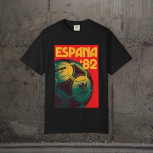 May include: Black t-shirt featuring a retro design with the text "ESPANA '82" in yellow and red. The design includes a soccer ball with the Adidas logo. The shirt is a classic style, suitable for casual wear.