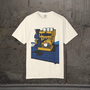 Op de afbeelding: Crèmekleurig T-shirt met een geel en blauwe illustratie van een espressomachine, een kopje koffie en een koffiemolen. Het ontwerp staat op een blauwe tafel. Het shirt heeft een ronde hals en korte mouwen.