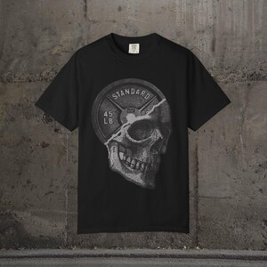 Może przedstawiać: Czarny t-shirt z grafiką talerza do podnoszenia ciężarów i czaszki. Na talerzu widnieje napis "STANDARD" i "45 LB." Czaszka jest częściowo widoczna, z szczegółowym cieniowaniem. Koszulka wykonana jest z miękkiego materiału.