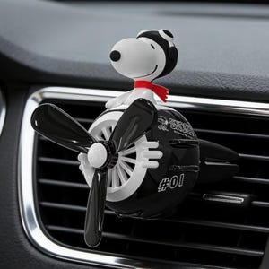 May include: A black and white Snoopy airplane car air freshener. The air freshener features a Snoopy figure wearing a pilot's hat and red scarf, sitting atop a propeller plane with the number "#01" on the side.