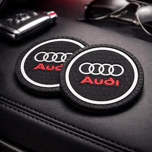 Może przedstawiać: Dwa czarne podkładki pod kubki do samochodu z logo Audi w kolorze białym i czerwonym. Podkładki mają białą obwódkę i teksturowaną powierzchnię. Logo Audi jest wyśrodkowane.