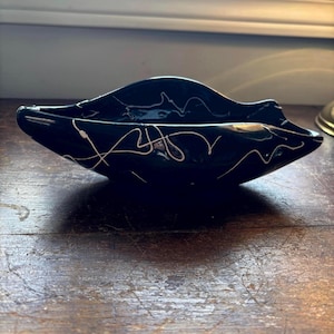 May include: A dark teal ceramic dish with an unusual, asymmetrical form. The dish has white, abstract squiggles across its surface. The bowl is displayed on a wooden surface, highlighting its glossy finish and decorative design.