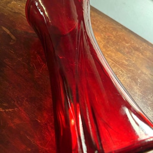 Peut inclure: Un vase en verre rouge, de forme élancée, avec une base évasée et un corps légèrement incurvé. Le vase a une finition brillante et reflète la lumière, créant des reflets et des ombres. Il est probablement utilisé pour les fleurs.