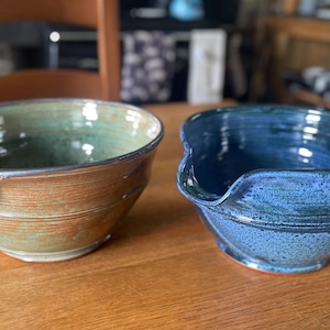 May include: Two ceramic mixing bowls with pouring spouts. One bowl features a variegated green and brown glaze with horizontal ridges, while the other bowl has a speckled deep blue glaze. Both bowls have a rounded shape and a smooth finish.