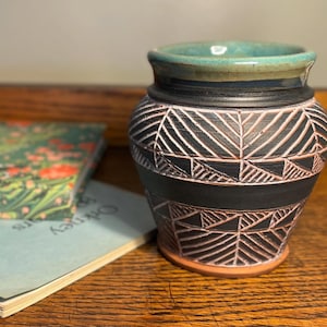 Puede incluir: Un jarrón de cerámica hecho a mano con borde turquesa, bandas negras y un patrón geométrico blanco. El jarrón está sobre una superficie de madera junto a un libro con diseño floral.