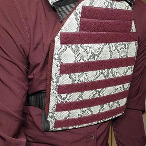 May include: A tactical vest with a snakeskin pattern and burgundy accents. The vest has multiple horizontal burgundy straps and a black adjustable strap. The vest is worn over a burgundy button-down shirt.