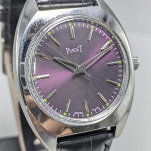 Puede incluir: Un reloj de pulsera Piaget plateado con esfera morada oscura y correa de cuero negro. La esfera del reloj tiene agujas e indicadores plateados, con el nombre de la marca. El reloj está marcado "Swiss Made".