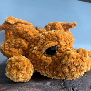 May include: A handmade, plush dragon in a warm, golden-orange hue. The crocheted dragon features a round body, small wings, and a detailed head with a black eye. The soft texture and whimsical design make it a charming decorative item.