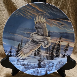 May include: A decorative plate featuring a snowy owl in flight over a winter landscape. The owl has white and gray feathers, with a backdrop of a sunset sky and snow-covered ground. The plate is displayed on a black stand.
