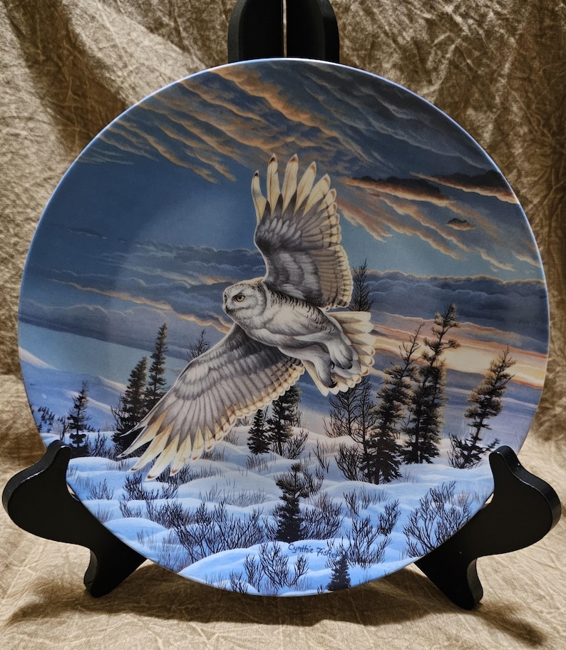 May include: A decorative plate featuring a snowy owl in flight over a winter landscape. The owl has white and gray feathers, with a backdrop of a sunset sky and snow-covered ground. The plate is displayed on a black stand.