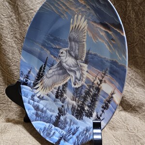 May include: Decorative plate featuring a snowy owl in flight over a winter landscape. The plate has a blue and white color scheme with a detailed painting of the owl, trees, and a snowy ground. The plate is displayed on a black stand.