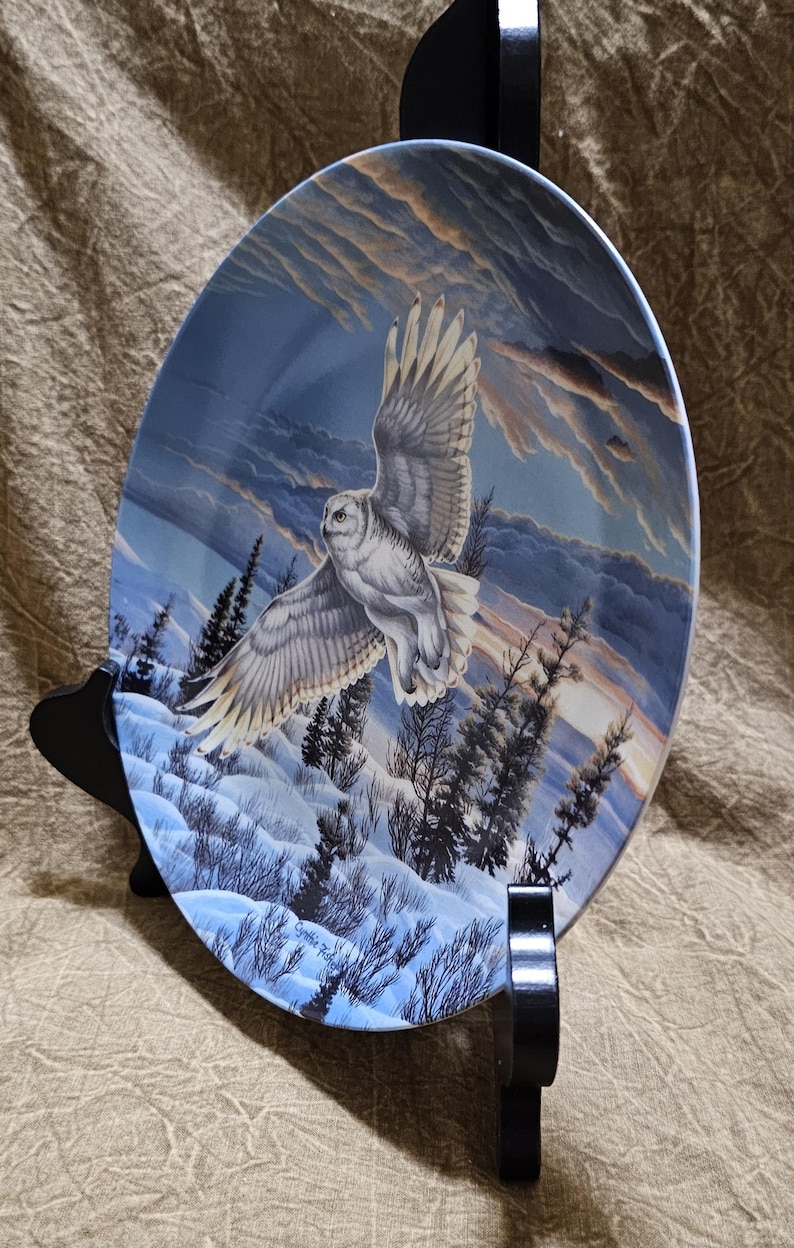 May include: Decorative plate featuring a snowy owl in flight over a winter landscape. The plate has a blue and white color scheme with a detailed painting of the owl, trees, and a snowy ground. The plate is displayed on a black stand.