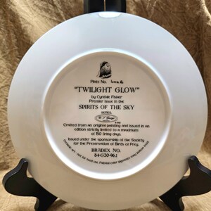 May include: A white ceramic plate with the text "TWILIGHT GLOW" by Cynthie Fisher, part of the "SPIRITS OF THE SKY" series. The plate is displayed on a black stand. The plate's text includes "BRADEX NO. 84-G20-96.1".