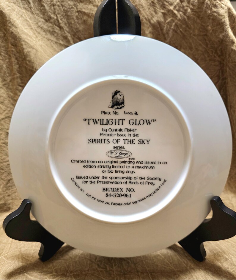 May include: A white ceramic plate with the text "TWILIGHT GLOW" by Cynthie Fisher, part of the "SPIRITS OF THE SKY" series. The plate is displayed on a black stand. The plate's text includes "BRADEX NO. 84-G20-96.1".