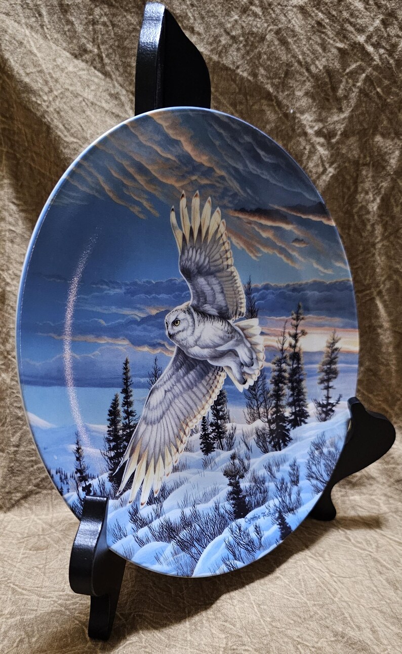 May include: Decorative plate featuring a snowy owl in flight over a winter landscape. The plate has a blue background with a painted scene of snow-covered ground, evergreen trees, and a cloudy sky. The plate is displayed on a black stand.