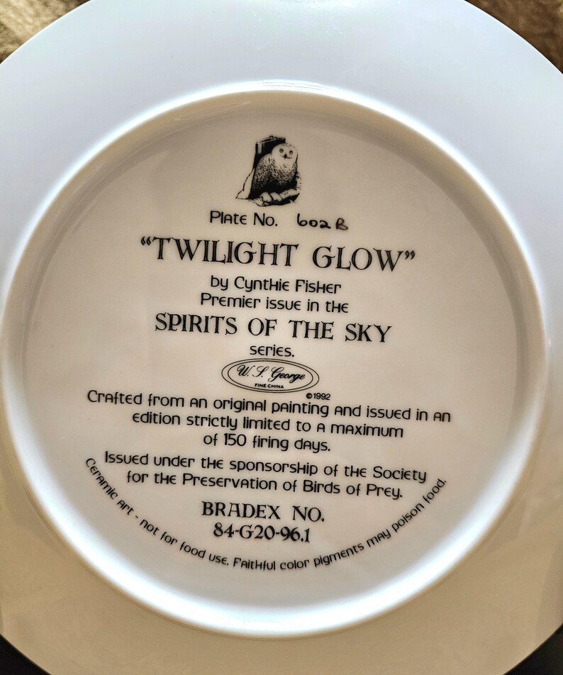 May include: A white ceramic plate with the text "TWILIGHT GLOW" and an illustration of an owl. The plate is part of the "SPIRITS OF THE SKY" series by Cynthia Fisher. It was issued under the sponsorship of the Society for the Preservation of Birds of Prey.