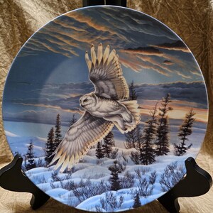 May include: Decorative plate featuring a snowy owl in flight over a winter landscape. The owl has white and gray feathers, with a backdrop of a sunset sky and snow-covered ground. The plate is displayed on a black stand.
