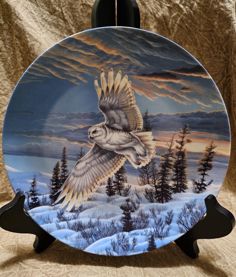May include: Decorative plate featuring a snowy owl in flight over a winter landscape. The owl has white and gray feathers, with a backdrop of a sunset sky and snow-covered ground. The plate is displayed on a black stand.
