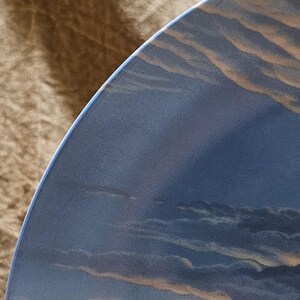 May include: A portion of a decorative plate featuring a painted landscape of a cloudy sky. The plate's edge is a deep blue, with the sky displaying shades of blue, gray, and tan. The design evokes a serene, natural scene.