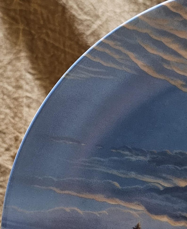 May include: A portion of a decorative plate featuring a painted landscape of a cloudy sky. The plate's edge is a deep blue, with the sky displaying shades of blue, gray, and tan. The design evokes a serene, natural scene.