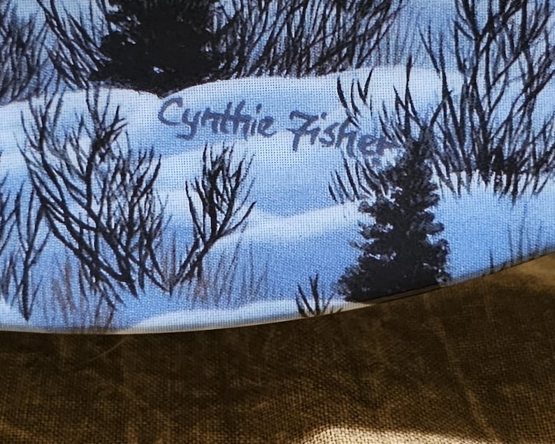 May include: A winter landscape painting featuring snow-covered ground, dark bare trees, and a small evergreen tree. The artist's signature, "Cynthia Fisher," is visible in the upper center. The color palette includes shades of blue, white, and black.