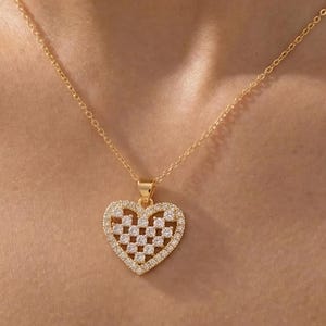 May include: A gold-colored necklace with a heart-shaped pendant. The pendant features a checkered pattern of clear, sparkling stones set within a gold frame. The necklace is displayed on a person's chest.