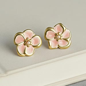 May include: A pair of flower-shaped stud earrings. Each earring features a pink enamel flower with a gold-colored border and a small pearl-like center. The earrings are displayed on a white surface.