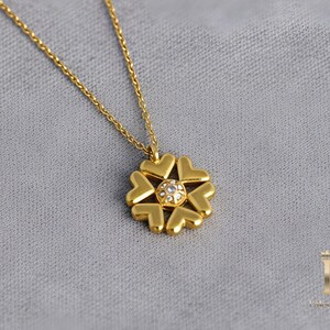 May include: A gold-colored necklace with a pendant shaped like a flower. The flower pendant is made of six heart-shaped petals surrounding a small, sparkling gem. The chain is delicate and gold, resting on a gray surface.