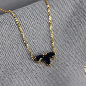 Puede incluir: Un delicado collar dorado con un colgante de tres gemas oscuras en forma de hoja. La fina cadena y el colgante crean un diseño minimalista. El collar descansa sobre un fondo de tela gris. Las gemas miden aproximadamente 1 cm.