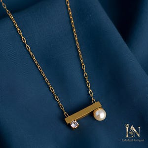 May include: A gold-colored necklace with a delicate chain and a horizontal bar pendant. The pendant features a small, clear gemstone and a white pearl. The necklace is displayed on a blue background.