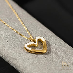 May include: Gold-tone necklace with a heart-shaped pendant. The pendant features a white enamel inlay on the edges, creating a contrasting design. The necklace is displayed on a gray surface, showcasing the jewelry's details.