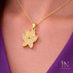 May include: A gold-colored butterfly pendant necklace. The pendant features a butterfly design with sparkling, clear stones. The necklace is on a gold chain. The background is a purple garment.