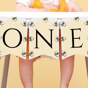 以下が含まれることがあります： 「ONE」と書かれたバナーが付いたハイチェア。クリーム色のバナーには黒い文字と蜂のイラストが描かれています。白いハイチェアには灰色の脚があり、赤ちゃんが座っています。
