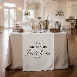 Puede incluir: Una mesa de recepción de bodas está decorada con una cubeta de champán, copas de flauta y una foto enmarcada. Un camino de mesa color crema muestra el texto "EST. 2025 MR. & MRS. Badesheim EST. 2025". La mesa está cubierta con un mantel de lino beige.