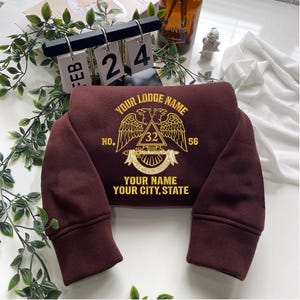 Puede incluir: Una sudadera granate con texto y gráficos bordados en dorado. El texto incluye "YOUR LODGE NAME", "NO. 32 56", "YOUR NAME" y "YOUR CITY, STATE". El diseño presenta dos águilas y una pancarta con las palabras "SPES MEA IN DEOES".