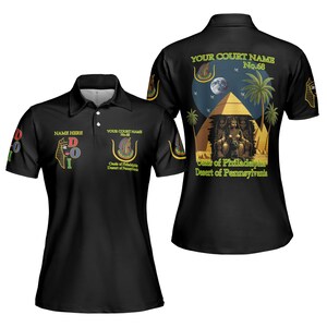 Könnte beinhalten: Schwarzes Poloshirt mit Kragen. Das Shirt zeigt verschiedene Grafiken, darunter ein ägyptisch inspiriertes Design mit Pyramiden, Palmen und dem Text "Oasis of Philadelphia Desert of Pennsylvania."