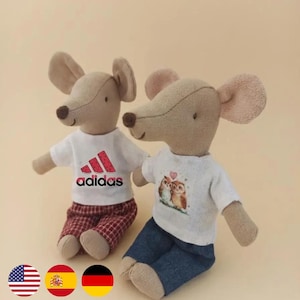 May include: Two beige fabric mouse dolls sitting side by side. The left mouse wears a white t-shirt with a watermelon-patterned Adidas logo and red plaid trousers. The right mouse wears a white t-shirt with an owl graphic and blue denim trousers.