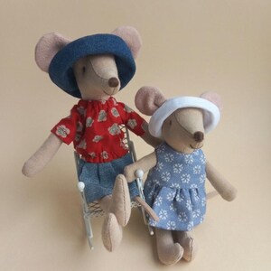 May include: Two beige fabric mouse dolls. The larger mouse wears a blue hat, a red floral shirt, and blue shorts, sitting on a white wire chair. The smaller mouse wears a white hat and a blue floral dress. Both have stitched facial features.
