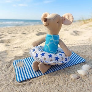 Op de afbeelding: Een beige stoffen muis-speeltje in een blauw badpak met een wit waterpatroon. De muis zit op een blauw-wit gestreepte stoffen mat en houdt een witte opblaasbare zwemband vast die is versierd met blauwe en grijze sterpatronen.