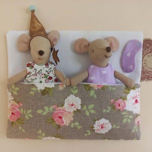 May include: Two plush mouse dolls tucked into a bed with a floral patterned blanket. One mouse wears a gold party hat and holds a star wand, while the other wears a purple dress with white hearts. A purple eye mask and a Maileg tag are visible.