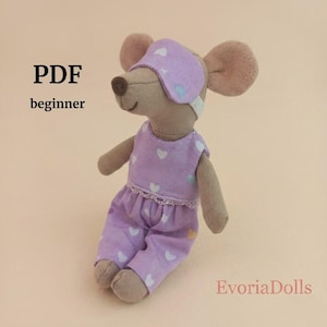 Peut inclure: Une poupée souris en tissu beige portant un masque de sommeil violet clair et une barboteuse assortie à motif de cœurs blancs. Le texte PDF beginner et EvoriaDolls est visible. La souris a de grandes oreilles et un petit nez marron.