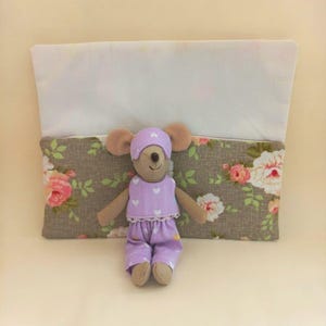 May include: A plush mouse toy wearing a light purple outfit with white hearts and a matching cap, positioned in front of a rectangular fabric pouch featuring a floral pattern of pink and white roses on a taupe background with a white top section.
