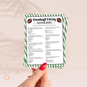 May include: A stack of green and white striped Football Trivia game cards. The cards feature the text "Football Trivia Halftime Shows" and questions about Super Bowl halftime performances. A hand with red nail polish holds the cards.
