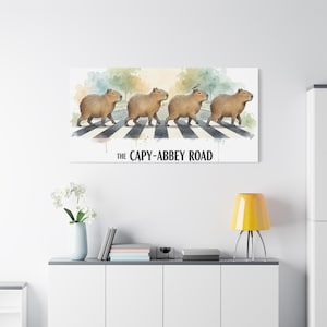 Könnte beinhalten: Leinwanddruck mit einer Aquarellillustration von Capybaras, die einen Zebrastreifen überqueren, mit einem weichen, farbenfrohen Hintergrund. Der Text "THE CAPY-ABBEY ROAD" ist unter dem Bild aufgedruckt. Das Kunstwerk ist eine humorvolle Anspielung auf das ikonische Beatles-Albumcover.