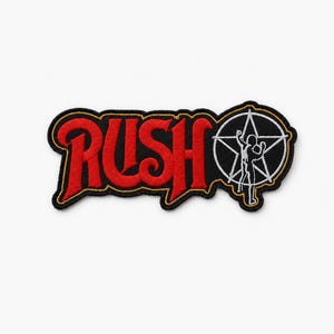 May include: An embroidered fabric patch featuring the word RUSH in bold red lettering with a black border. To the right of the text is a white star-man emblem enclosed within a white circle, all set against a black background with gold trim.