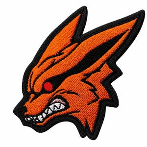 May include: An embroidered patch featuring the stylised head of a fox with bright orange fur, a black outline, and sharp pointed ears. The fox has a fierce expression with a red eye, a snarling mouth, and visible white teeth against a black background.