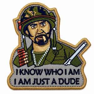 May include: An embroidered patch featuring a soldier in a camouflage helmet and green uniform, smoking a cigar, with a bandolier and rifle. Below the figure, the text reads: I KNOW WHO I AM I AM JUST A DUDE. The patch has a tan border and gray background.
