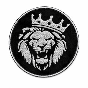 Pode incluir: Um emblema bordado circular apresentando uma cabeça de leão branca com uma expressão rugidora e uma coroa na cabeça, sobre um fundo preto sólido com uma borda costurada na cor prata ao redor de toda a borda circular do emblema.